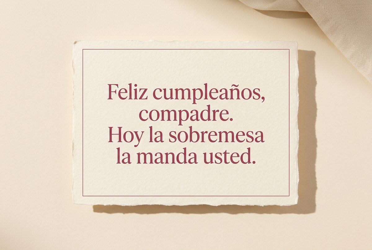 Tarjeta de cumpleaños de papel crema hecho a mano, con borde deckle y un filete impreso en rosa oscuro, fotografiada en plano cenital. En la tarjeta se lee «Feliz cumpleaños, compadre. Hoy la sobremesa la manda usted.». Al lado, una servilleta de tela crema doblada en la esquina superior derecha.