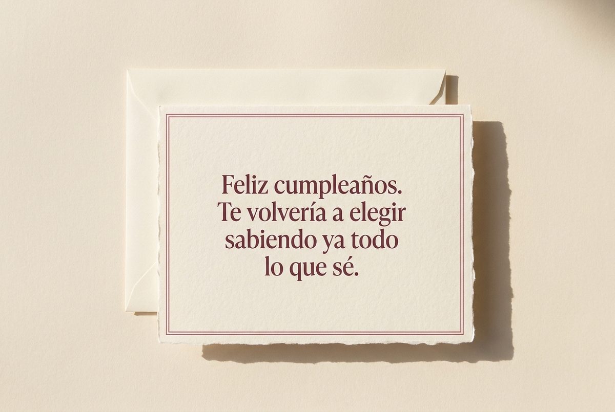 Tarjeta de cumpleaños de papel crema hecho a mano, con borde deckle y un filete impreso en rosa oscuro, fotografiada en plano cenital. En la tarjeta se lee «Feliz cumpleaños. Te volvería a elegir sabiendo ya todo lo que sé.». Al lado, un sobre crema abierto por detrás de la tarjeta.