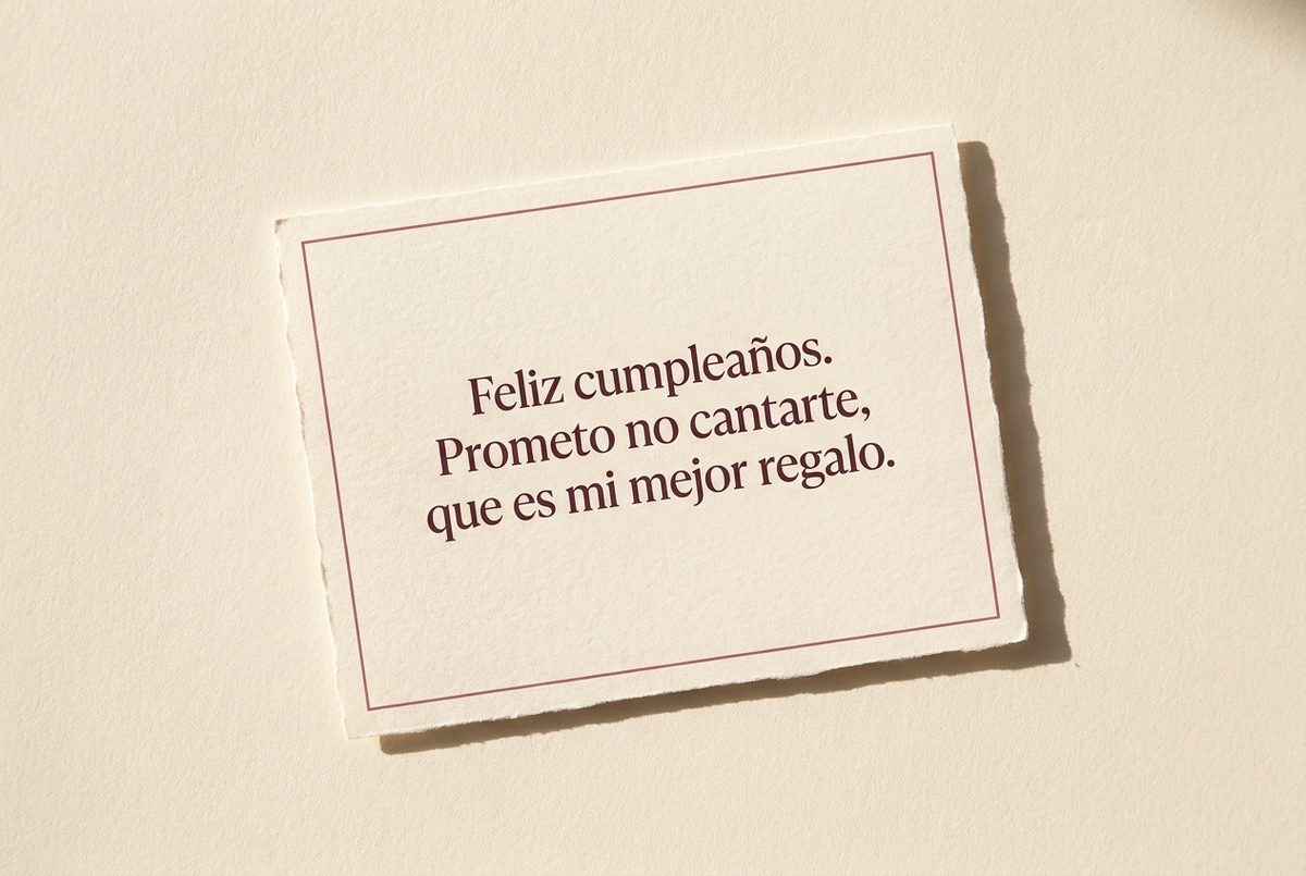 Tarjeta de cumpleaños de papel crema hecho a mano, con borde deckle y un filete impreso en rosa oscuro, fotografiada en plano cenital. En la tarjeta se lee «Feliz cumpleaños. Prometo no cantarte, que es mi mejor regalo.». Al lado, la tarjeta ligeramente ladeada, sin ningún otro objeto.