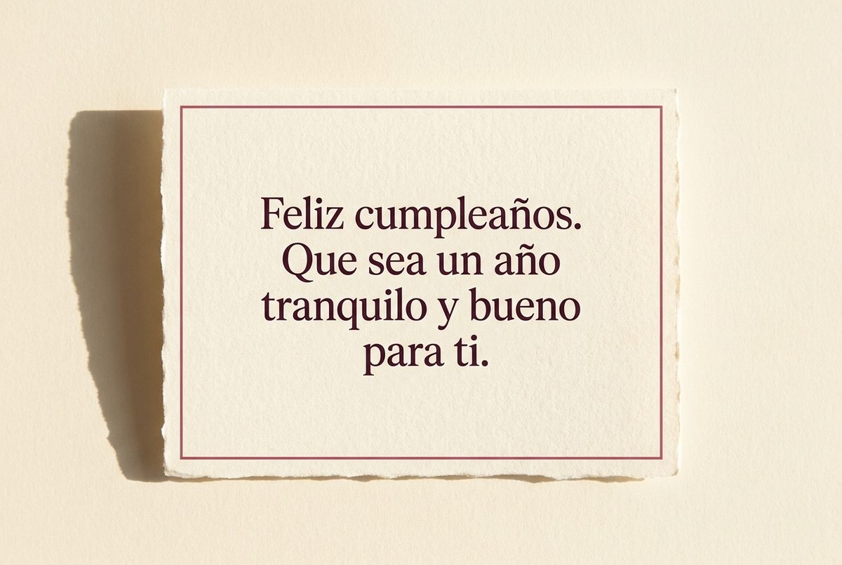 Tarjeta de cumpleaños de papel crema hecho a mano, con borde deckle y un filete impreso en rosa oscuro, fotografiada en plano cenital. En la tarjeta se lee «Feliz cumpleaños. Que sea un año tranquilo y bueno para ti.». Al lado, la tarjeta sola, con una sombra larga a la derecha.